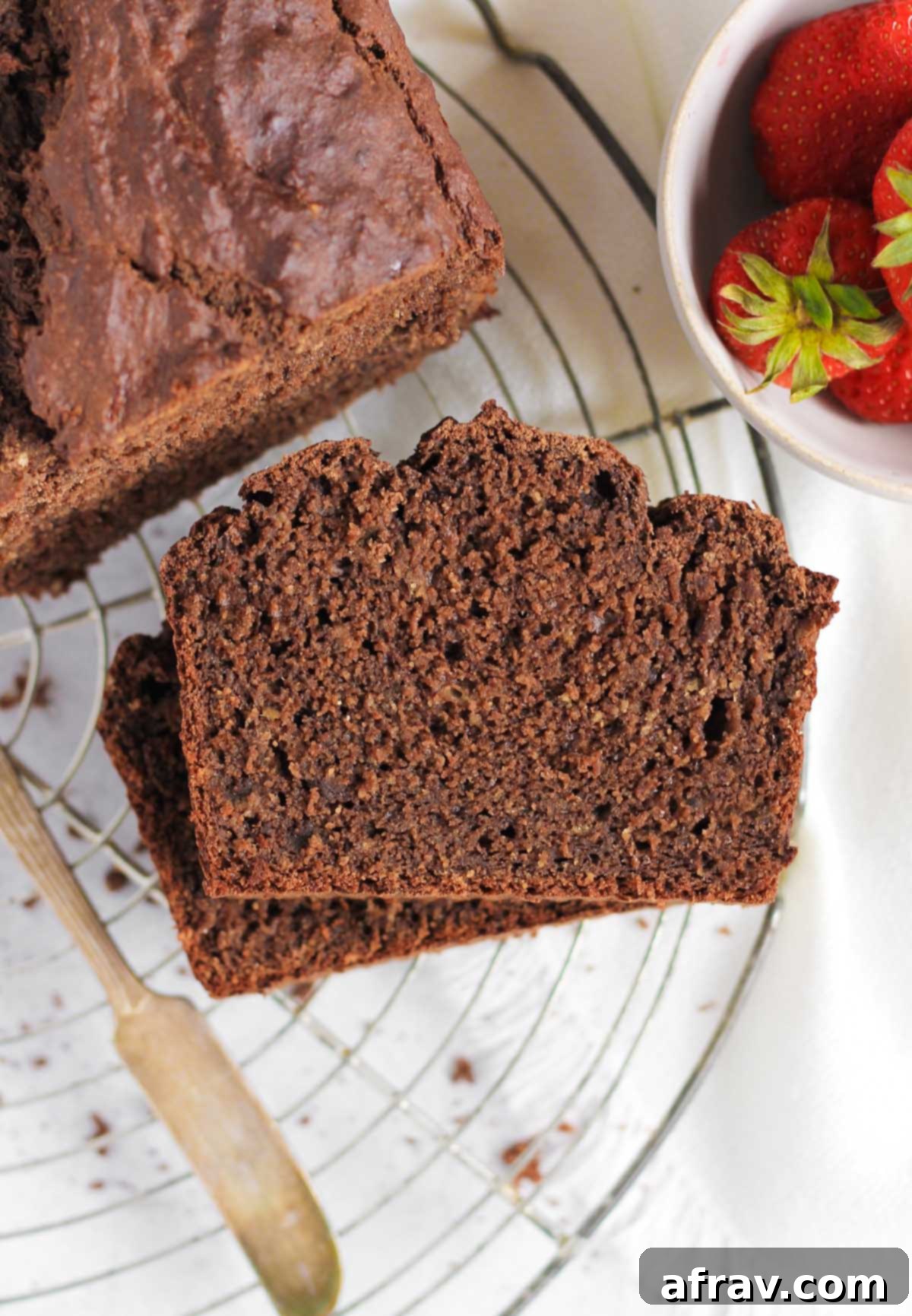 Two slices stacked of chocolate protein banana bread on a round wire cooling rack with the full loaf and a bowl of strawberries to the side.