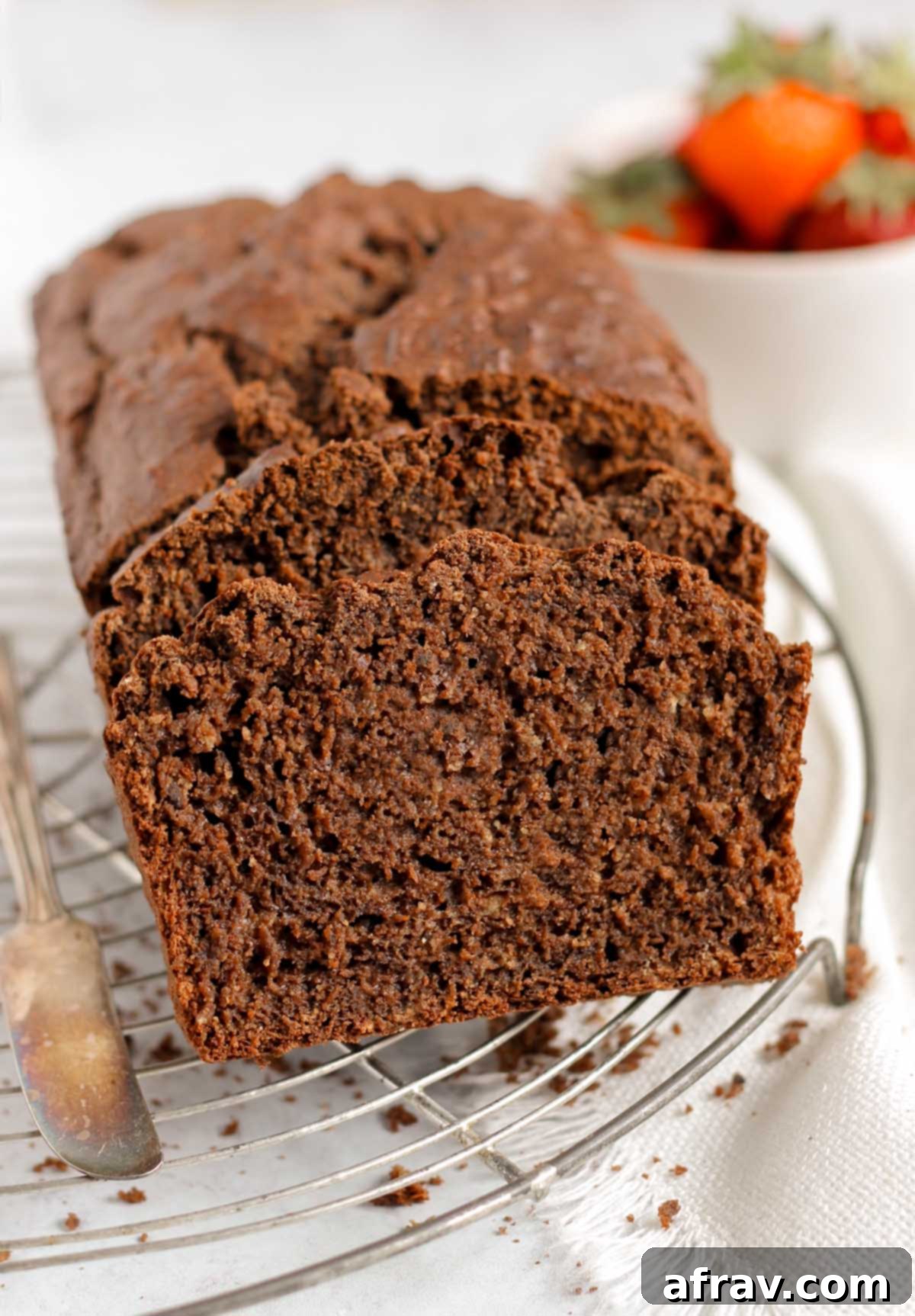 Loaf of chocolate protein banana bread partially sliced on a wire cooling rack with strawberries in the background.