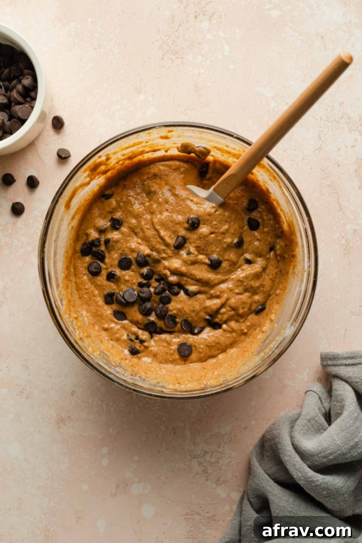 Pumpkin cake batter in a glass bowl topped with chocolate chips and stirred with a spatula, showing its rich color and consistency.