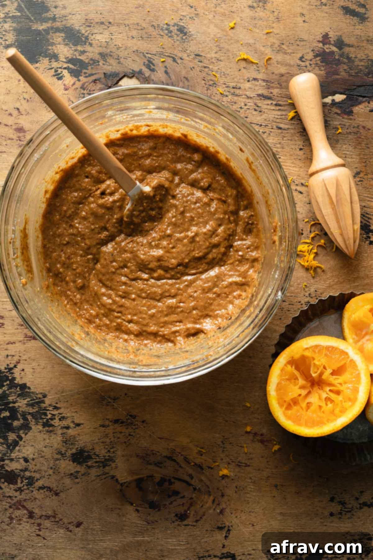 Orange spice cake batter in a glass bowl with a spatula stirring it, showing its smooth, light brown consistency.