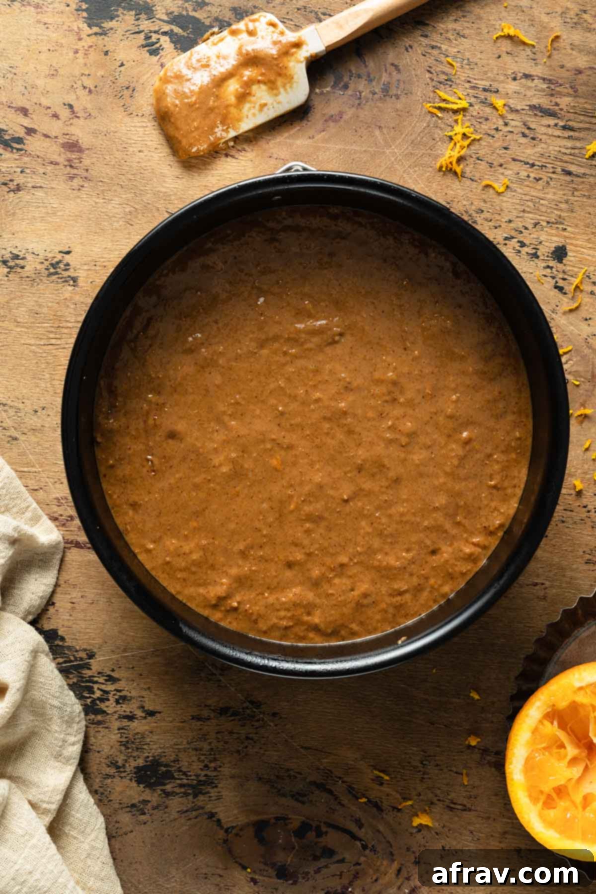 Smooth orange spice cake batter evenly distributed in a round black cake pan, ready for the oven.