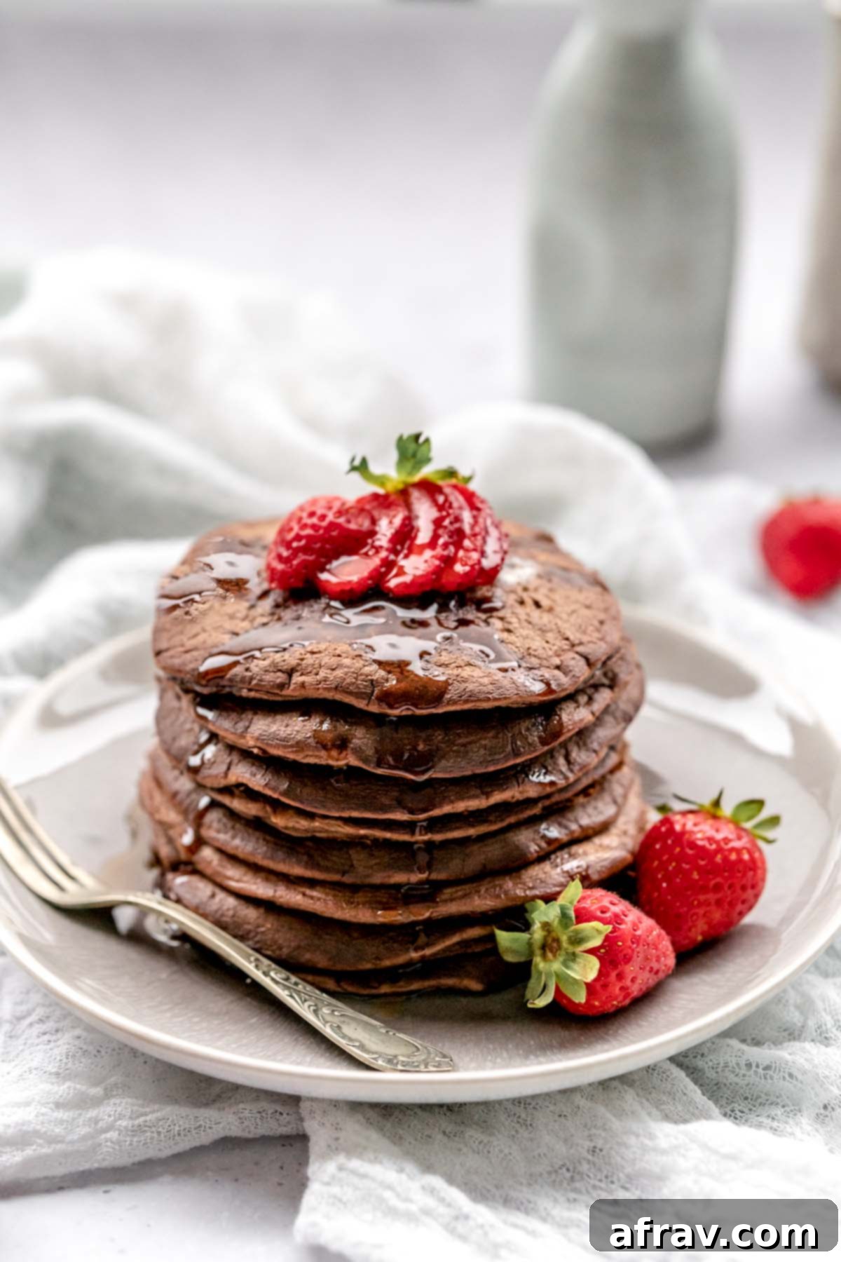 Chocolate pancakes topped with strawberries and dripping with syrup on a plate with a vintage fork.