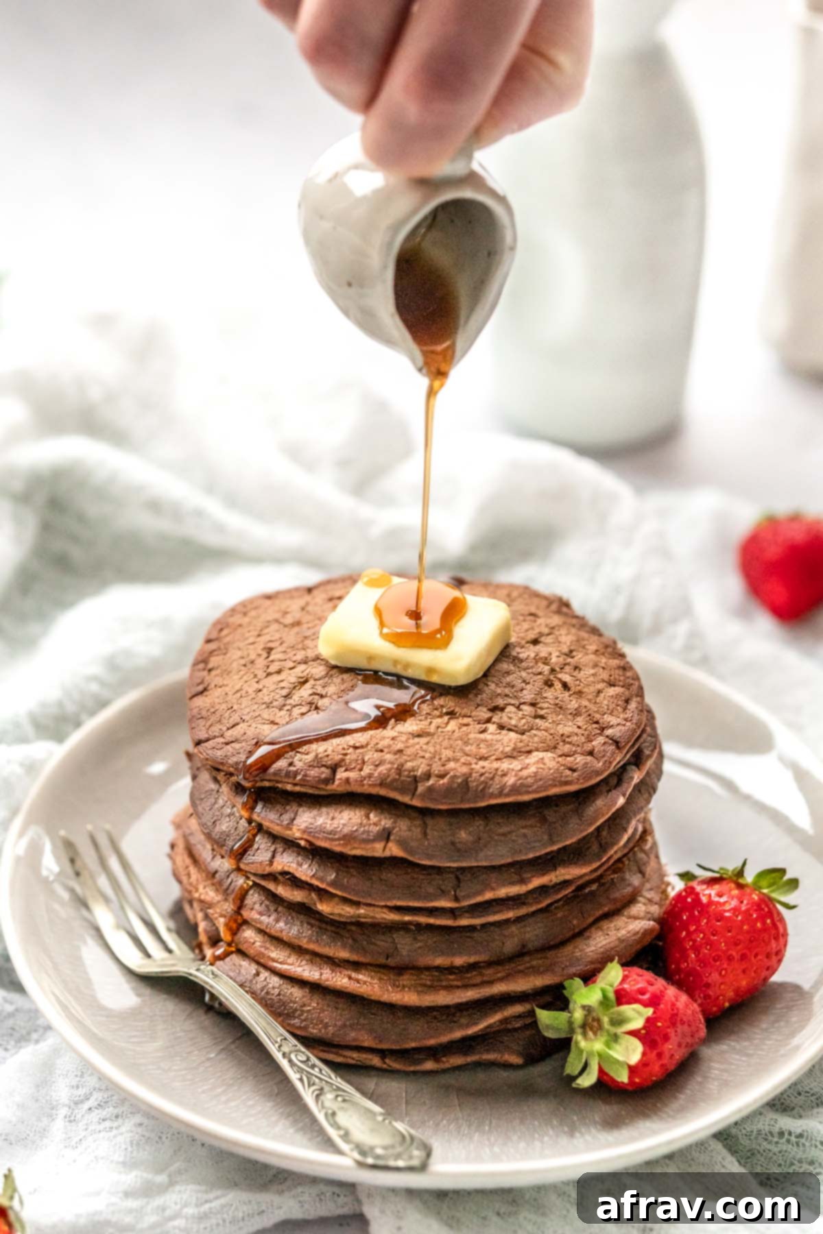 Syrup being poured over a stack of chocolate pancakes topped with a butter square and strawberries on the side.