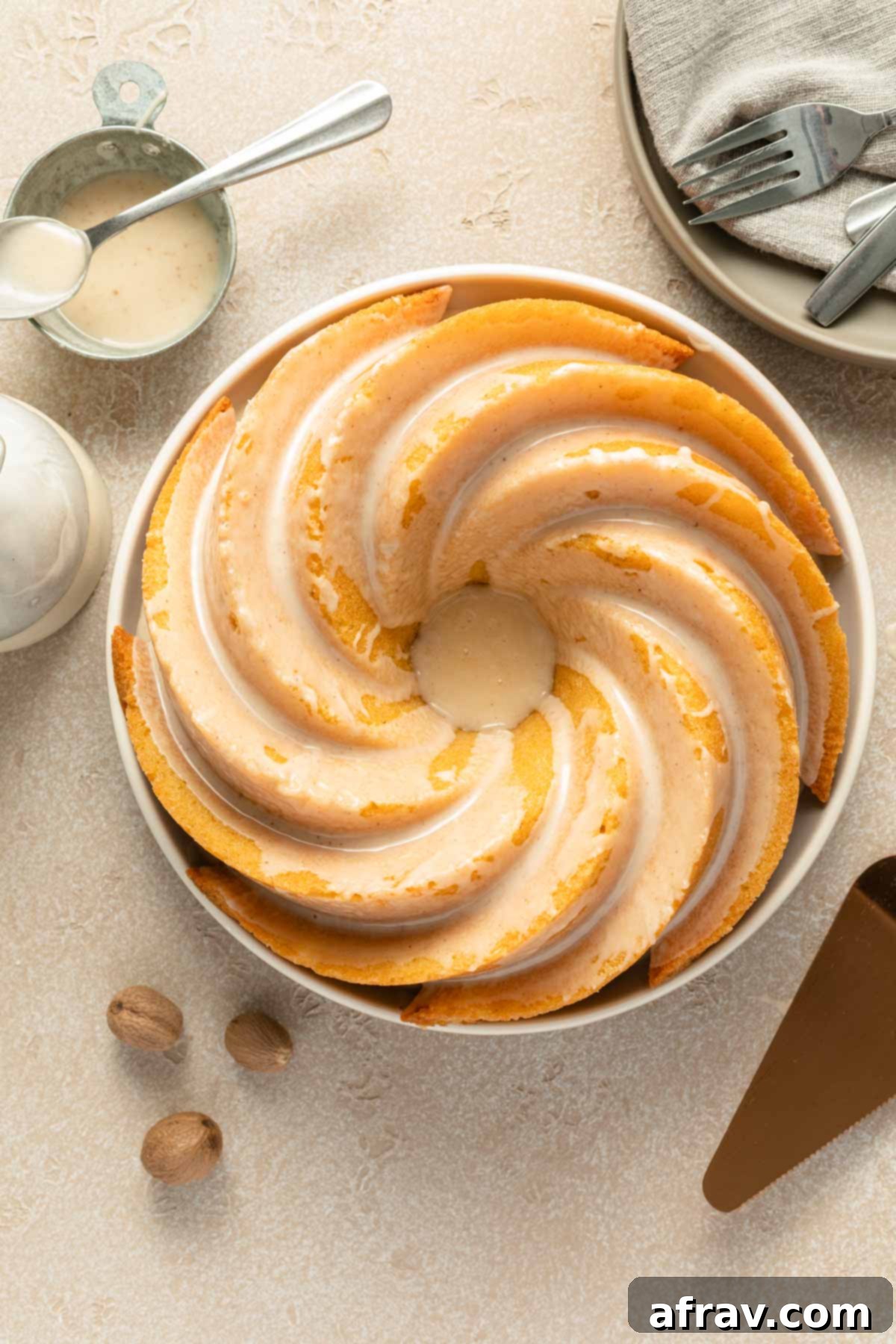 Overhead view of a perfectly baked and glazed Eggnog Bundt Cake on a decorative plate, ready to be served for the holidays. The golden-brown cake is generously coated with a white, drippy glaze.