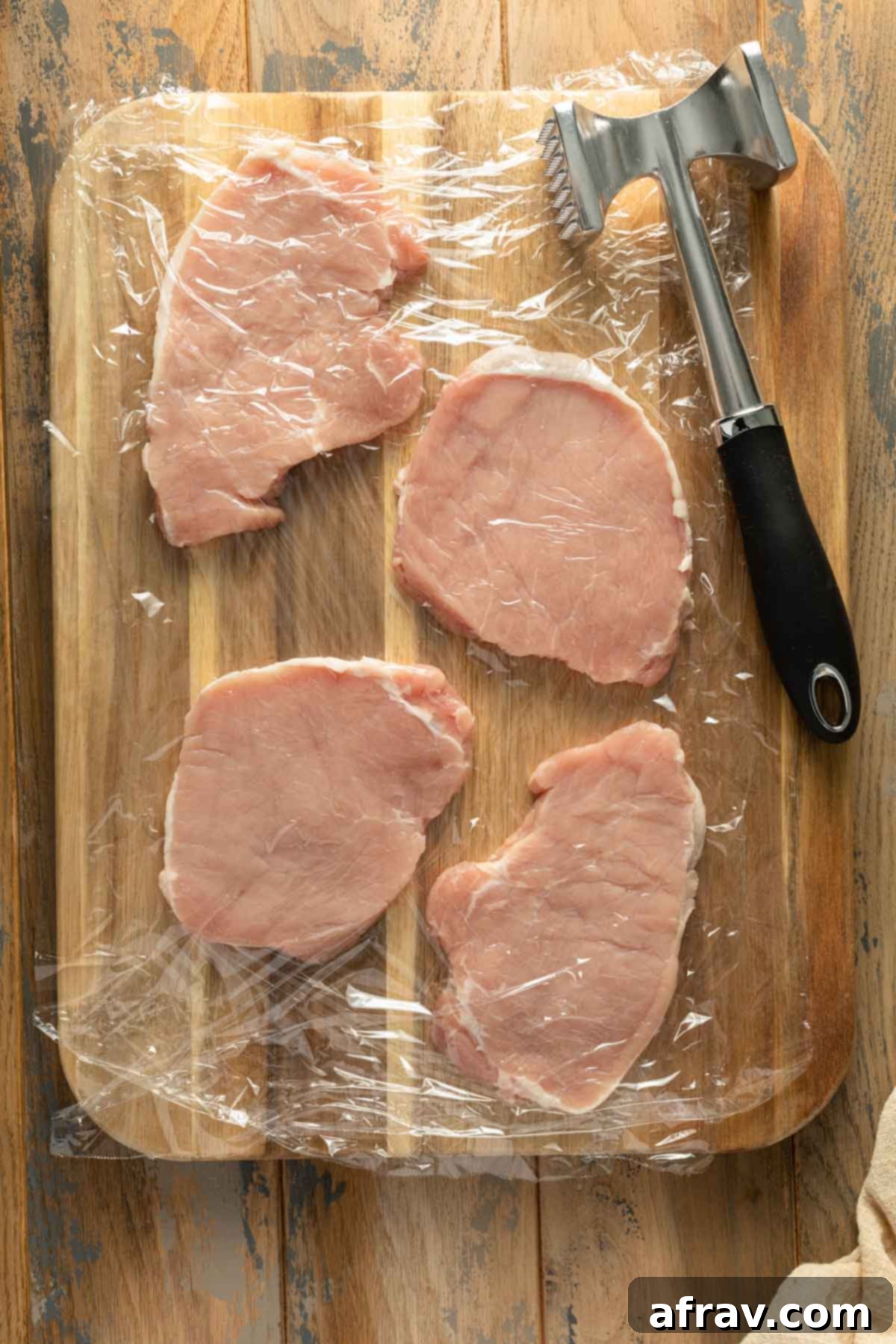Pork chops on a wooden board, covered with plastic wrap, next to a meat mallet for pounding.
