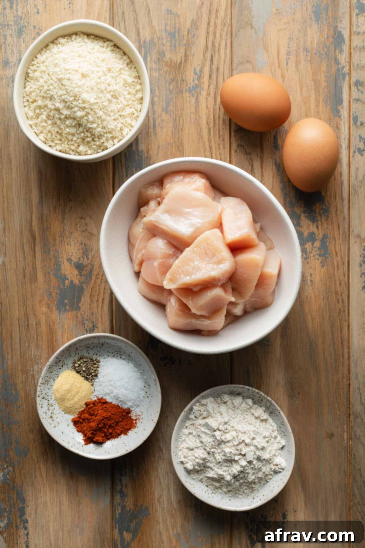 Individual ingredients for breaded chicken bites, including chicken, flour, eggs, panko, and spices, laid out on a wooden table.