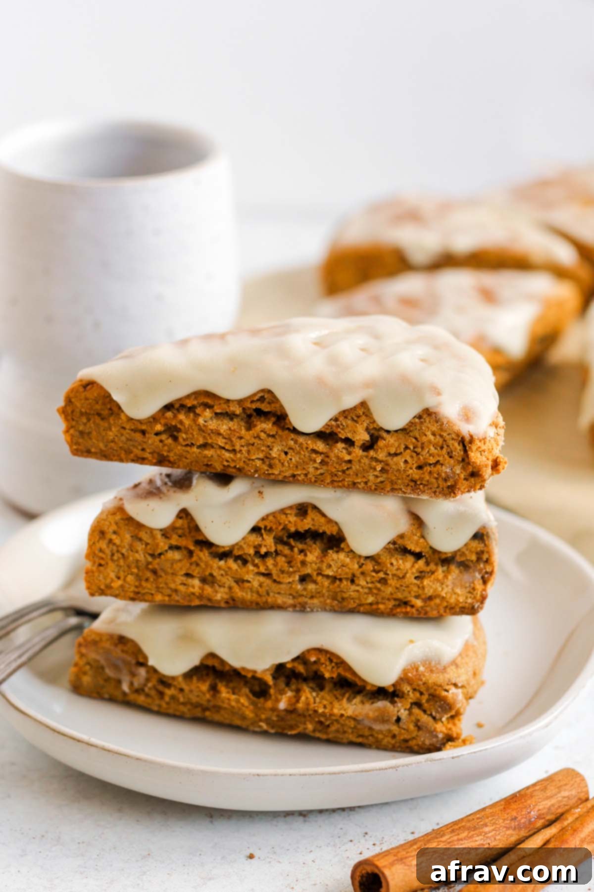 Pumpkin scones stacked 3 high on a plate with a ceramic mug and additional scones in the background.