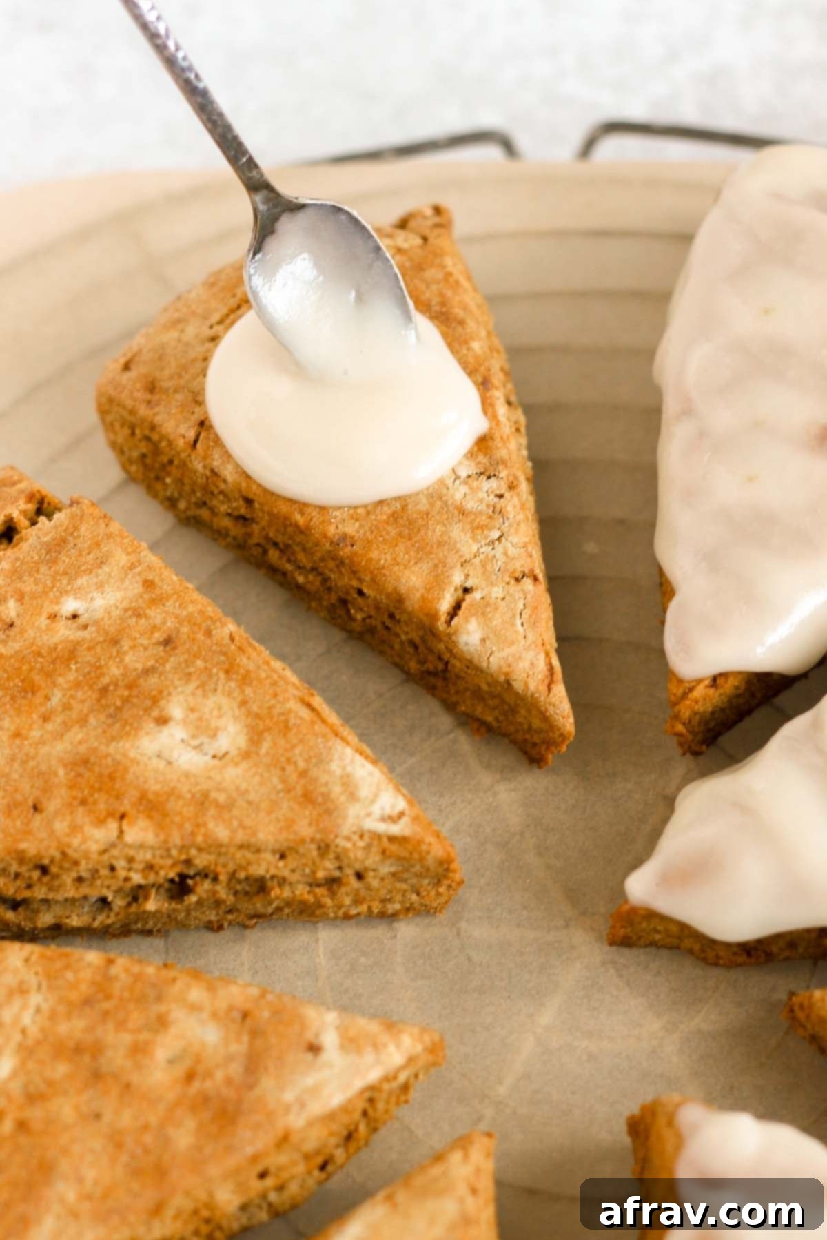 A spoon dripping a white glaze over baked scones on a round cooling rack.