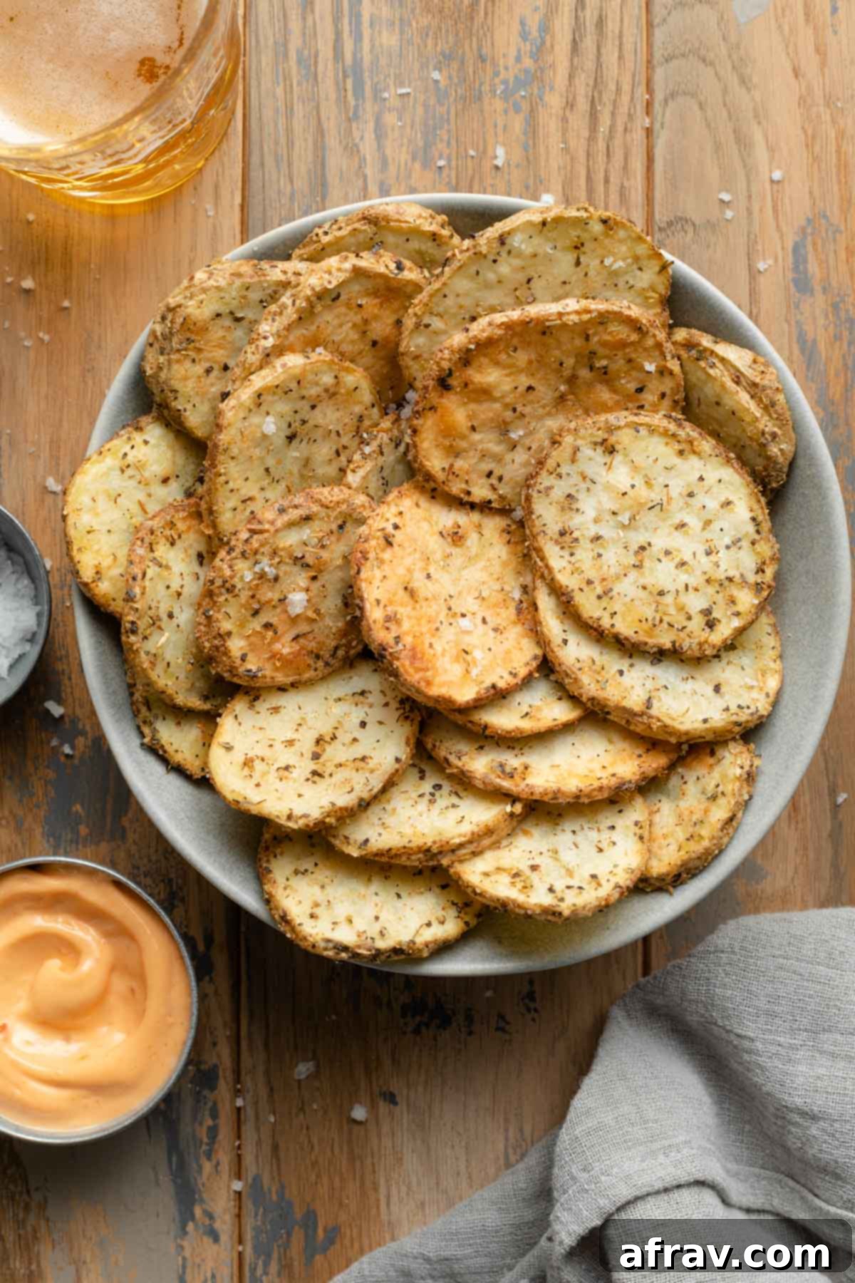 Overhead view of air fryer potato slices in a bowl on wooden backdrop next to dip, a drink and a grey napkin.