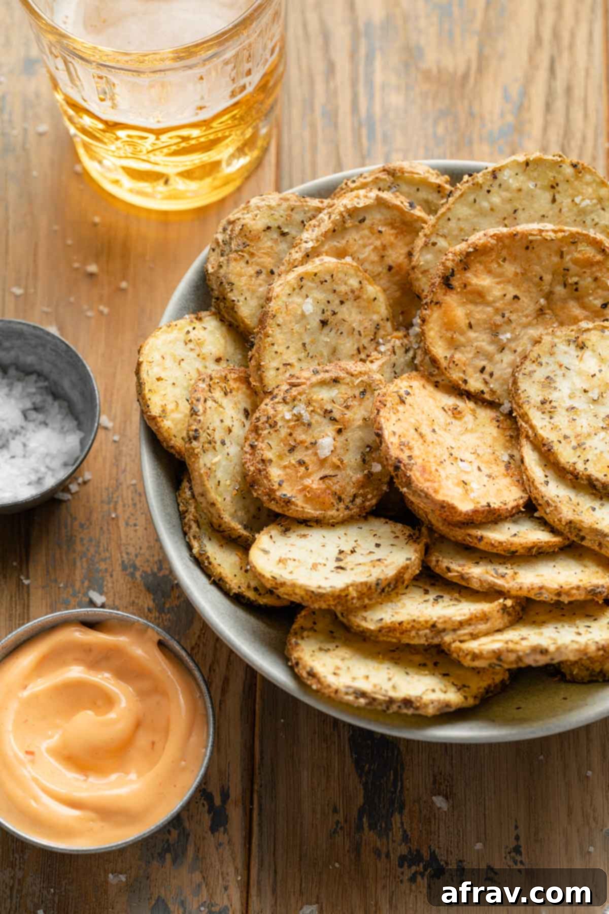 Angled view of potato slices in a grey bowl next to a bowl of dip and a drink in the background.