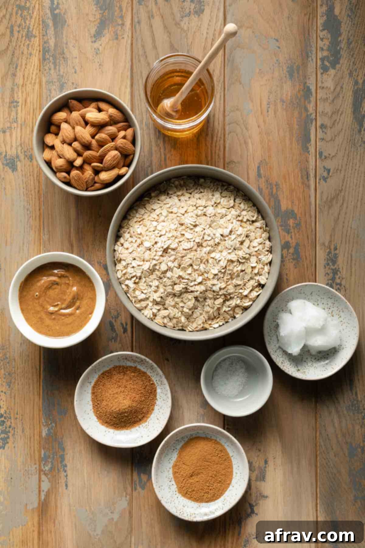 Ingredients to make honey almond granola arranged individually on a wooden table surface including oats, almonds, honey, coconut oil, cinnamon, coconut sugar, almond butter, and salt.