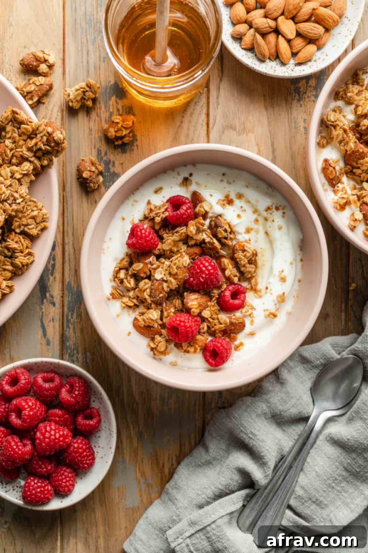A bowl of yogurt, raspberries, and honey almond granola on a wooden table, with fresh raspberries, a honey dipper, and granola scattered nearby.
