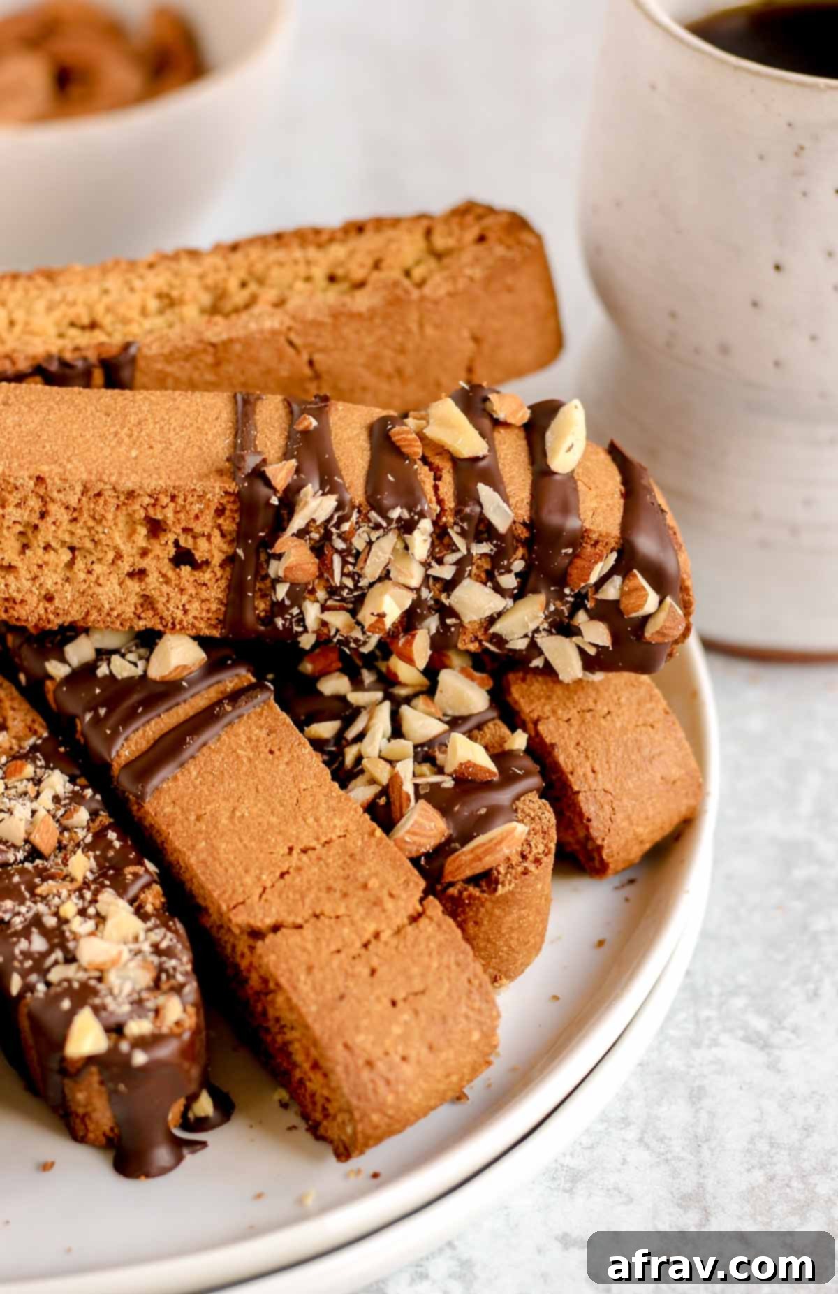 Biscotti drizzled with chocolate and almond pieces stacked on a plate with a cup of coffee in the background.