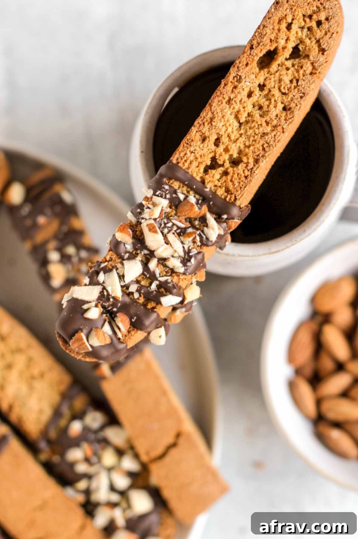 Single biscotti drizzled with chocolate and almond pieces laying onto of a cup of coffee with additional biscotti in the background.