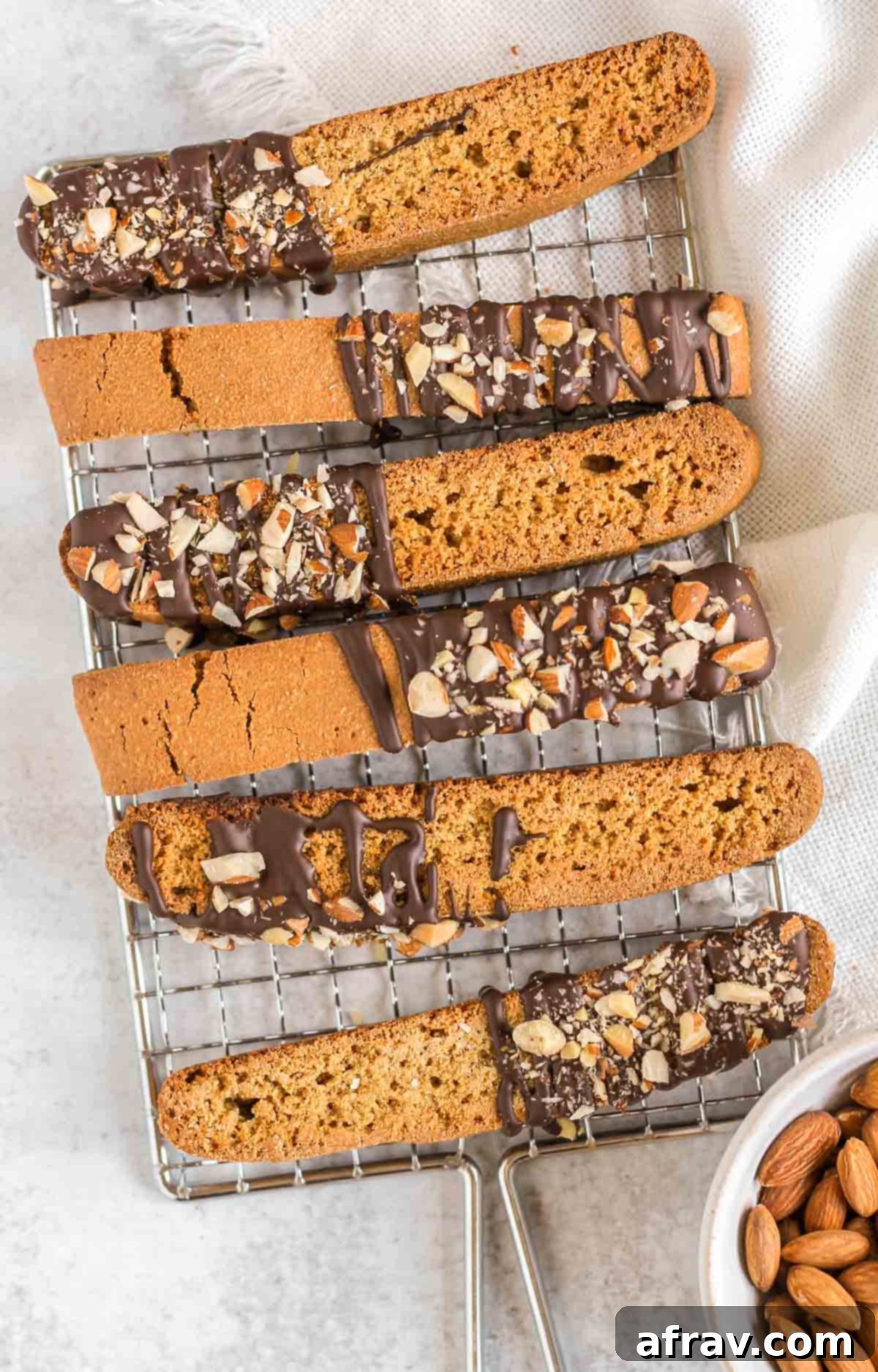 Biscotti cookies drizzled with chocolate and topped with chopped almonds on a small drying rack with a bowl of almonds on the side.