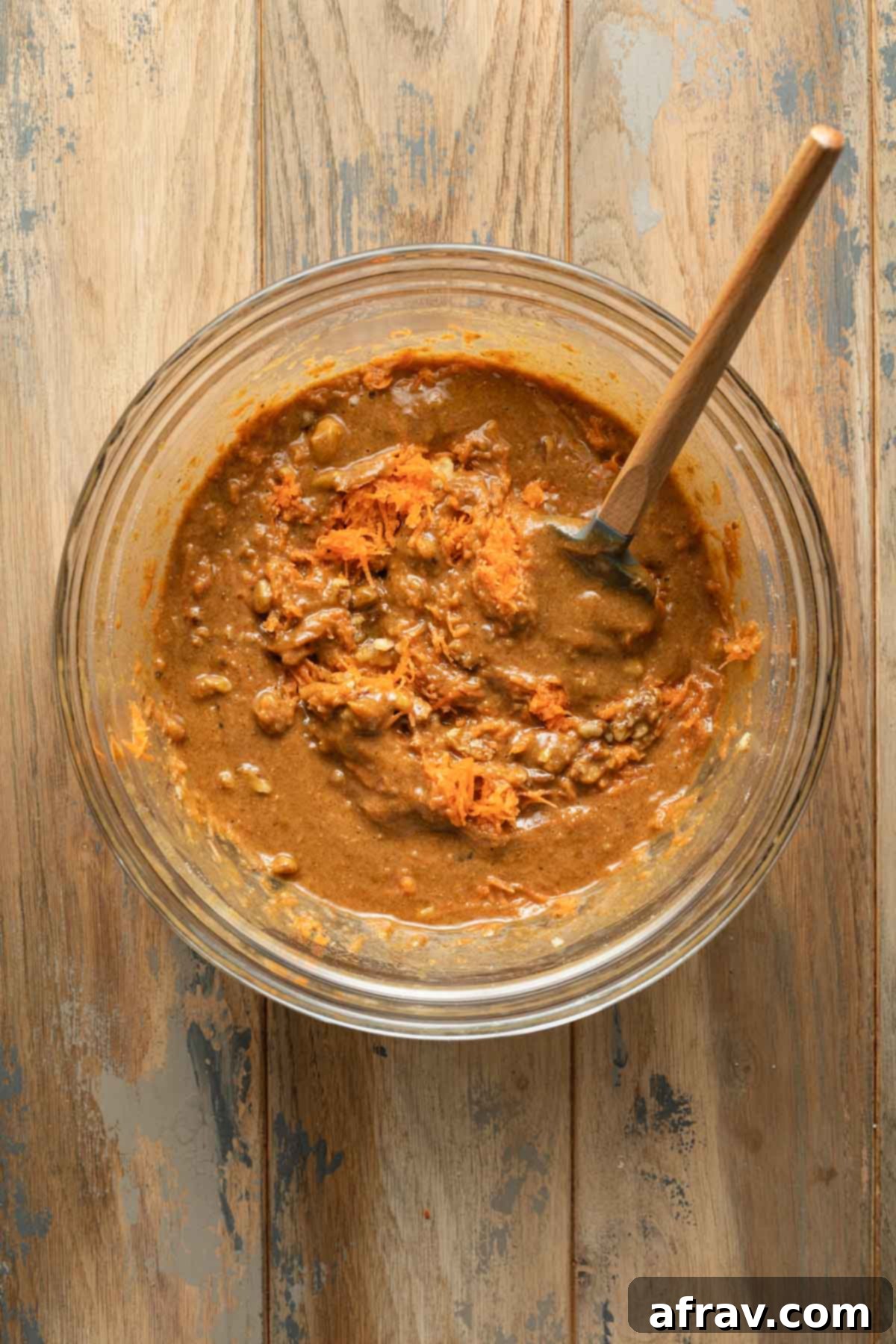 Close-up of carrot cake batter in a glass bowl, with shredded carrots and chopped walnuts visibly mixed in using a spatula, showing the rich texture.
