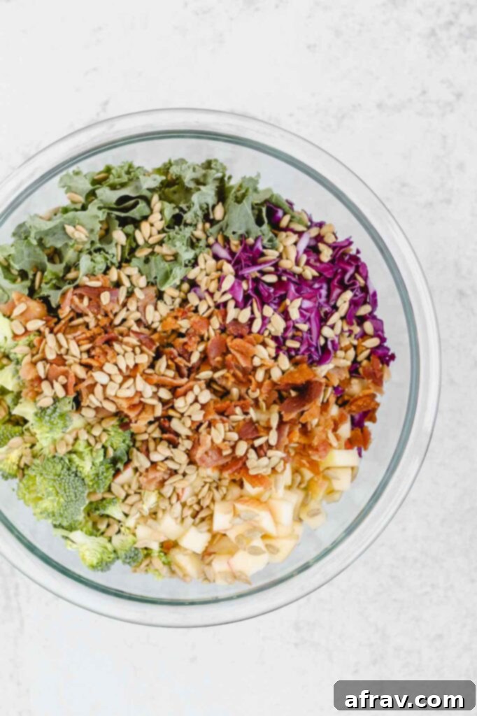 Broccoli, kale, bacon, cabbage, apples, and sunflower seeds gathered in a clear glass mixing bowl, ready for dressing.