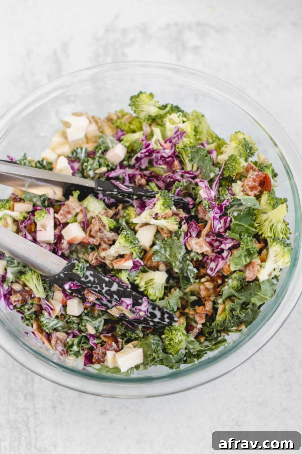 Close-up of tongs mixing the broccoli kale salad with the creamy dressing in a large bowl.