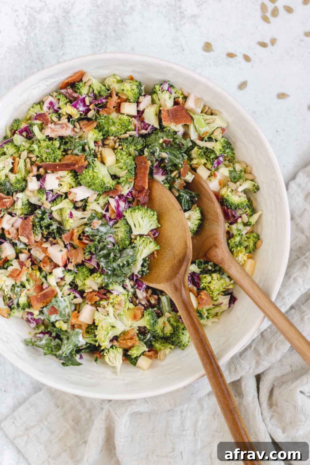 Broccoli kale salad in a beautiful white bowl with wooden salad serving utensils.