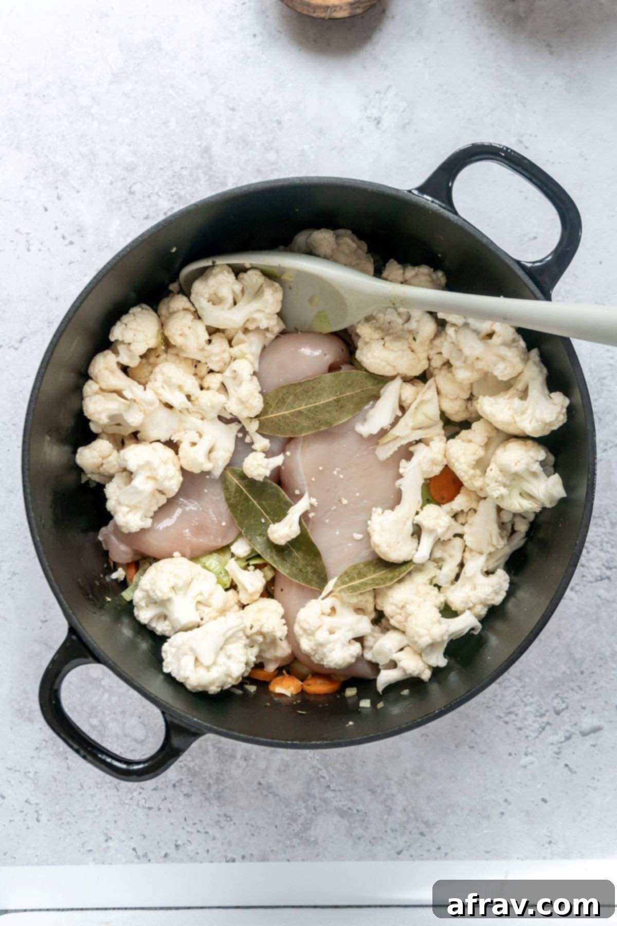 Veggies, raw chicken breasts, cauliflower florets, and bay leaves in a large black stock pot.
