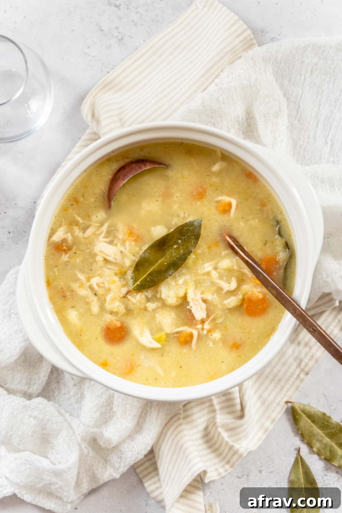 A white soup crock filled with creamy chicken and cauliflower soup with a bay leaf laying on top and a copper soup ladle in the soup.