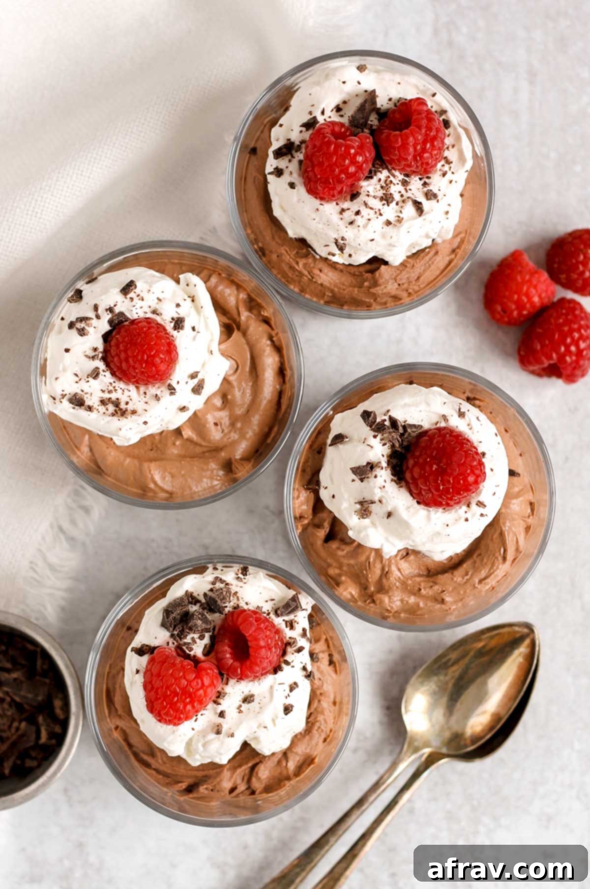 Four singe serve glass dishes filled with vegan chocolate mousse and topped with whipped cream, raspberries, and chocolate shavings.