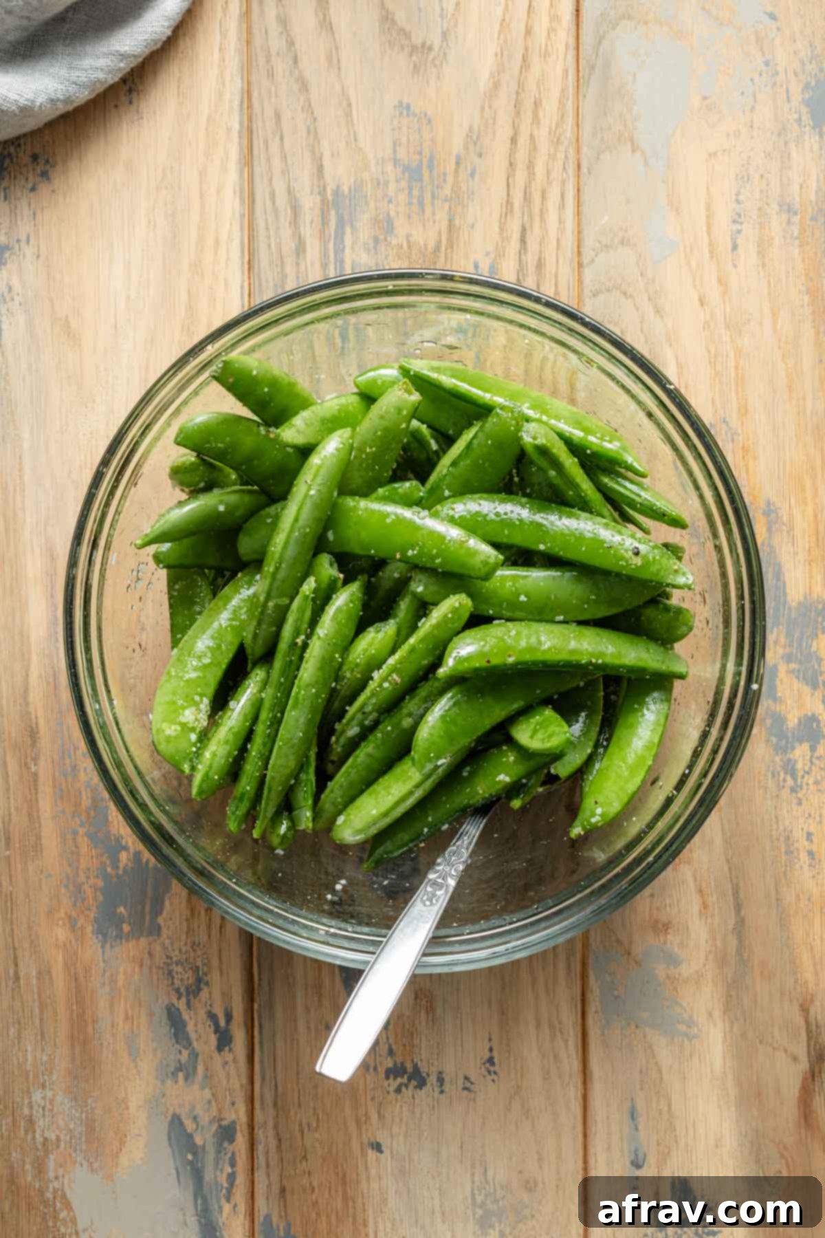 Crispy Air Fryer Snap Peas 4 Fresh sugar snap peas coated in olive oil and a mix of salt, pepper, and garlic powder, ready for air frying in a clear glass bowl, showcasing the seasoning process.