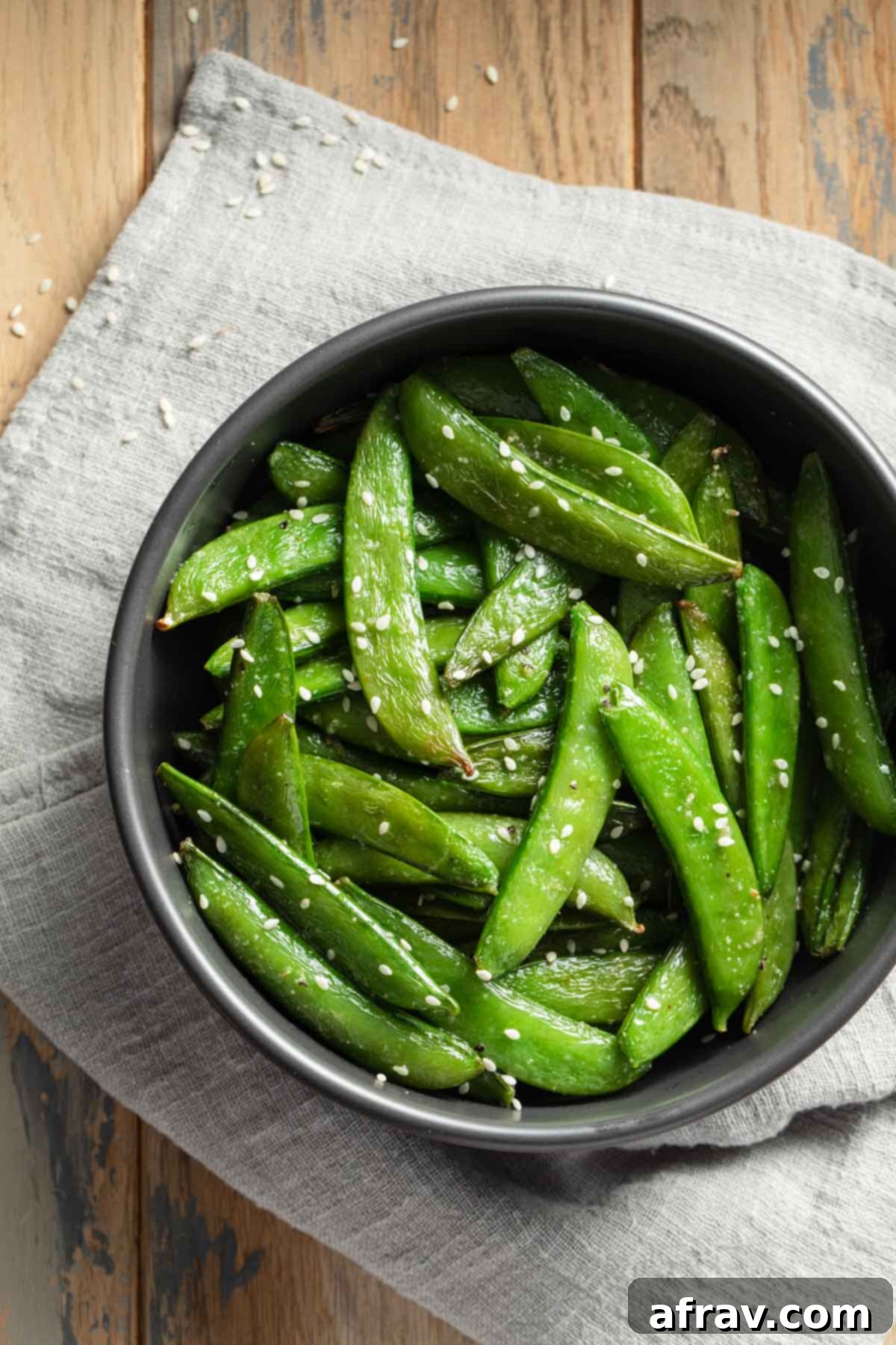 Crispy Air Fryer Snap Peas 7 A black bowl filled with perfectly roasted sugar snap peas, garnished lightly with sesame seeds, resting on a gray linen napkin, highlighting their inviting presentation as a healthy side.