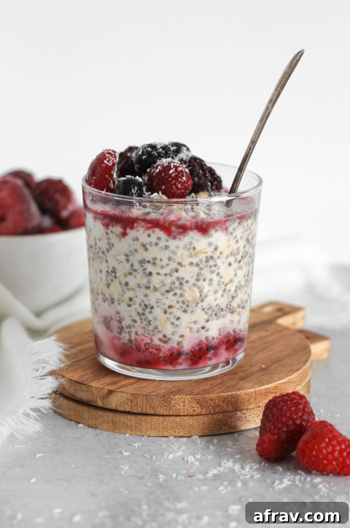 Glass cup, on a small round wooden cutting board, filled with overnight oats topped with thawed frozen berries with a vintage spoon in the glass.