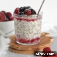 Glass cup, on a small round wooden cutting board, filled with overnight oats topped with thawed frozen fruit with a vintage spoon in the glass.