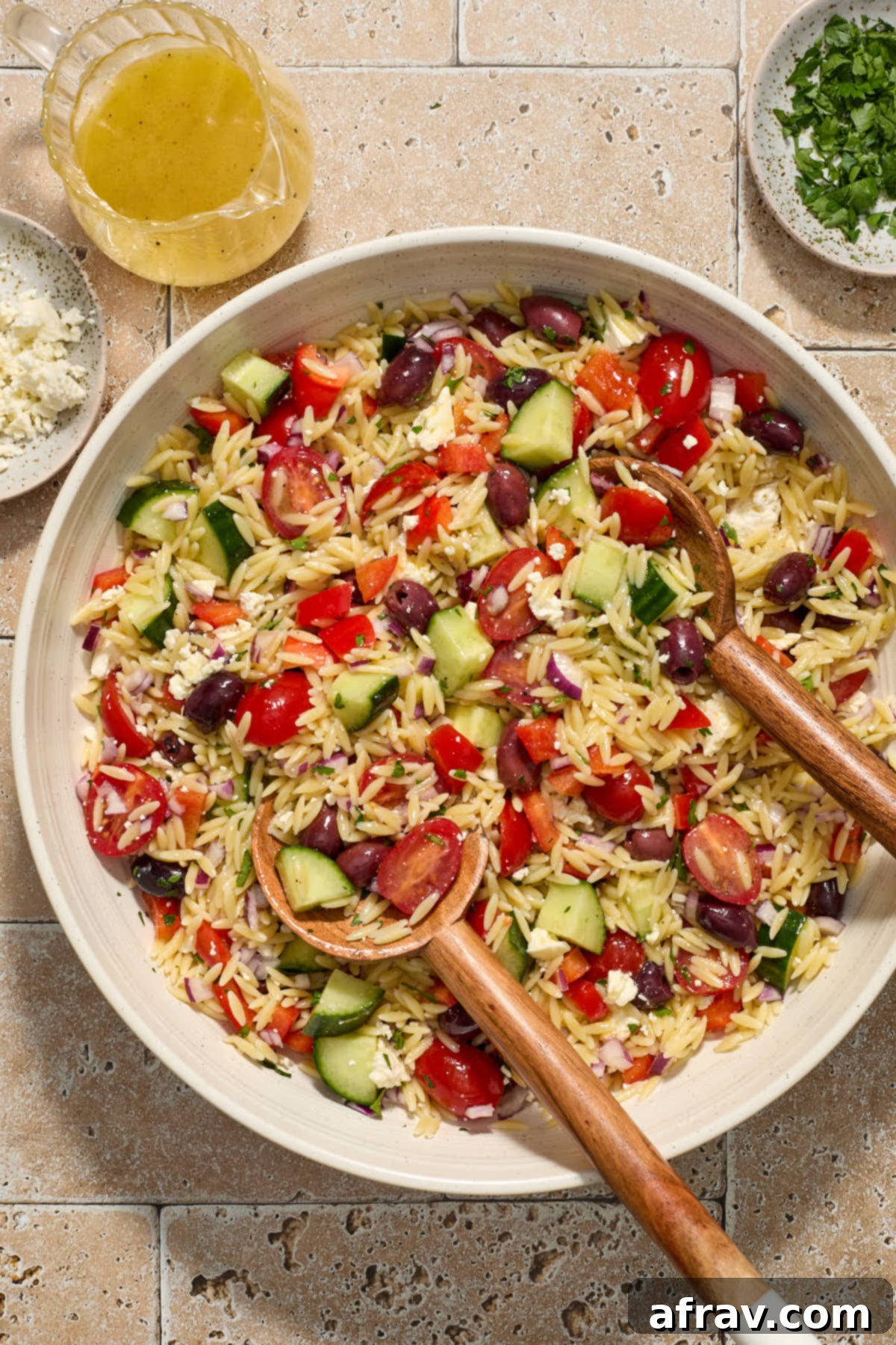 A large white bowl filled with vibrant Greek orzo pasta salad, featuring tender orzo, chopped vegetables, feta, and olives. Wooden salad servers are placed in the bowl, with fresh parsley, crumbled feta, and lemon slices scattered around, along with a small jug of lemon dressing.
