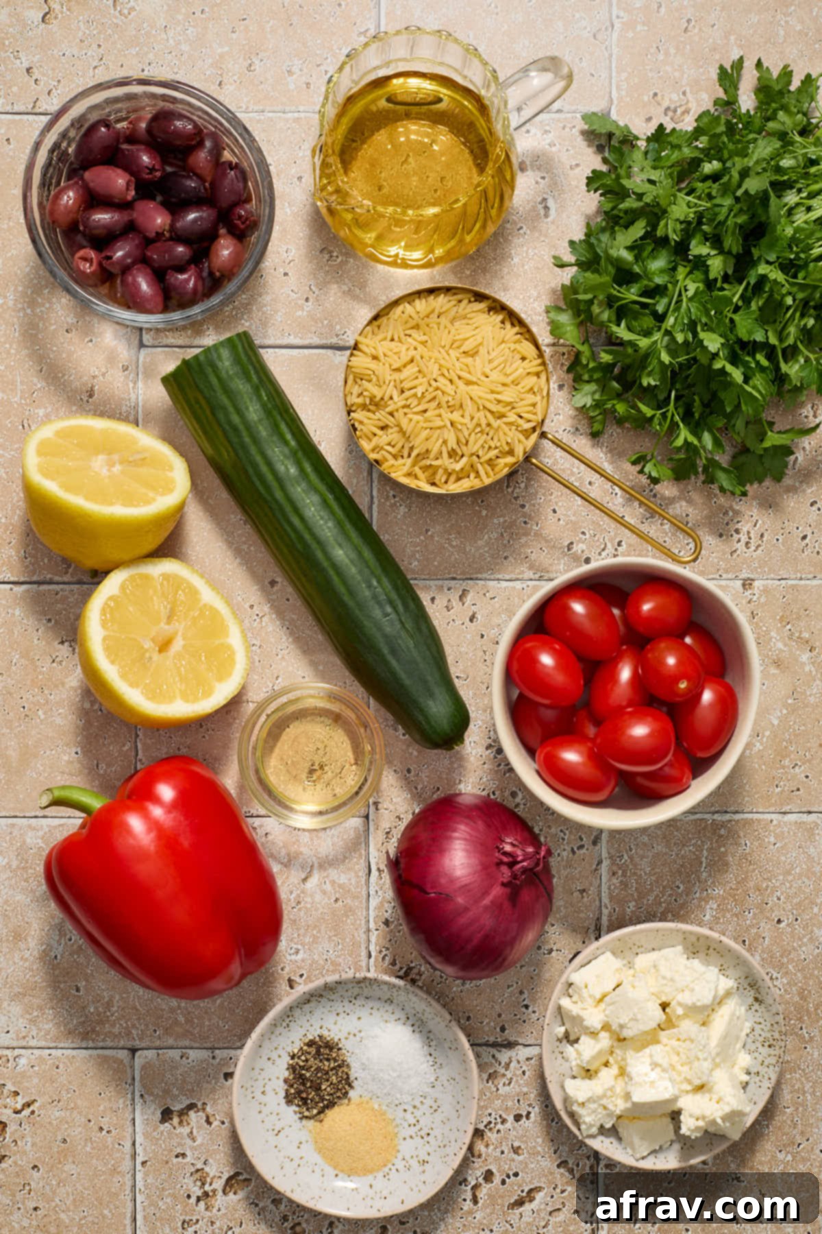Individual ingredients for Greek orzo salad arranged on a white surface, including uncooked orzo, chopped cucumber, bell pepper, tomatoes, red onion, pitted Kalamata olives, crumbled feta, fresh parsley, and a small jug of lemon vinaigrette ingredients.
