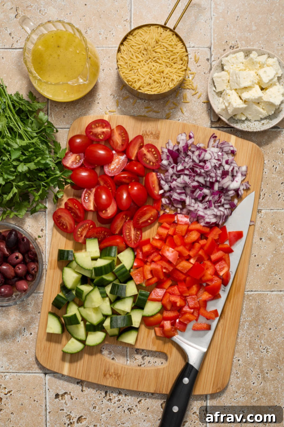 Assorted chopped vegetables including cucumber, bell pepper, tomatoes, red onion, and fresh parsley, arranged on a wooden cutting board with a knife nearby. Other salad ingredients like olives and feta are in small bowls around the board.