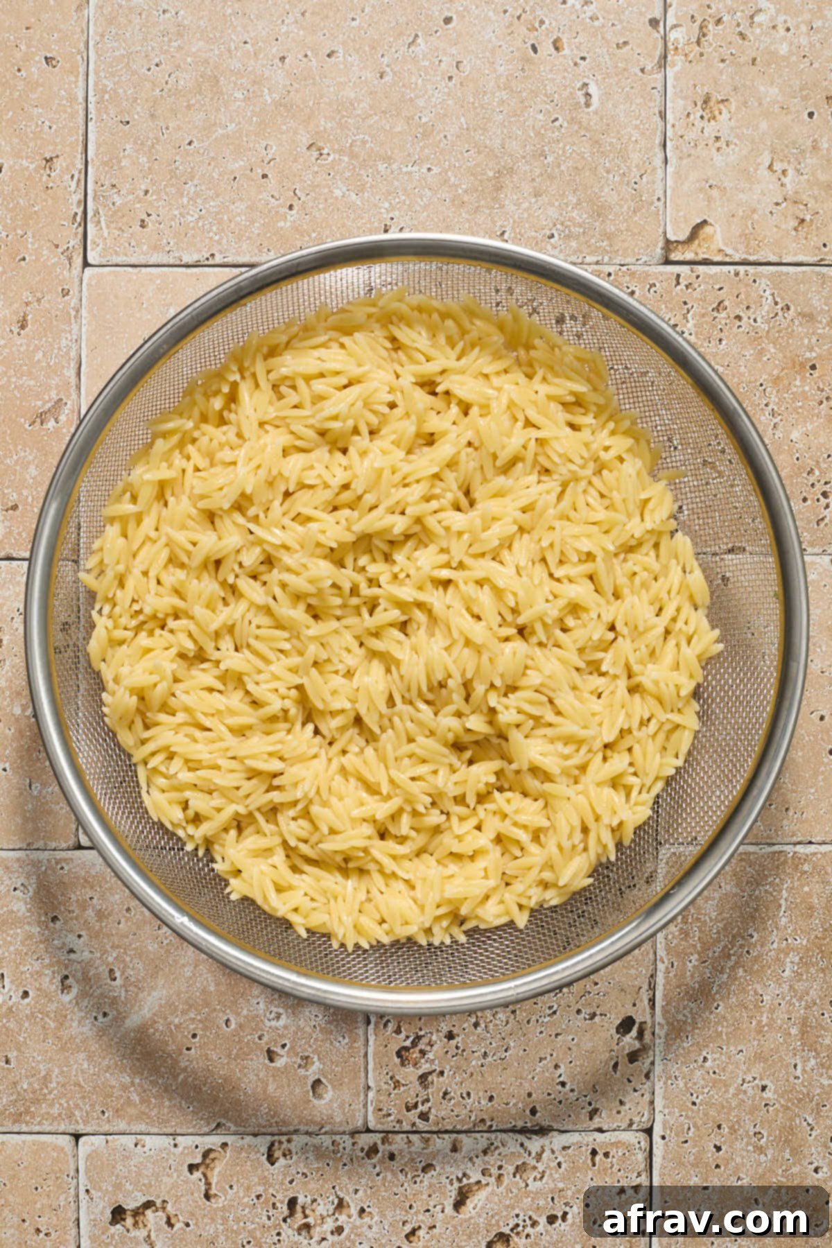 Cooked orzo pasta being rinsed under cold water in a mesh strainer, with steam rising.