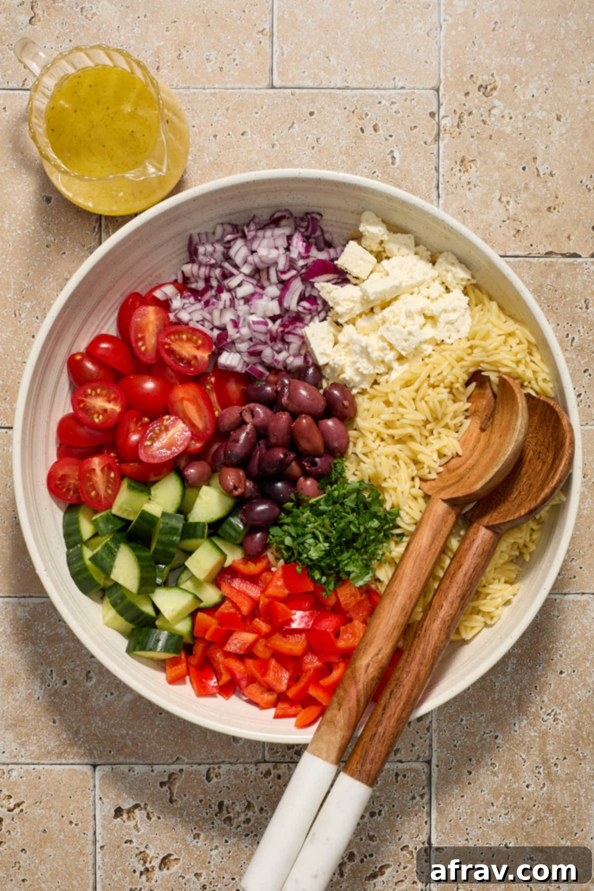 A large white bowl with cooked orzo pasta at the bottom, topped with chopped vegetables, olives, and crumbled feta, ready for the vinaigrette to be added. Salad servers are resting next to the bowl.