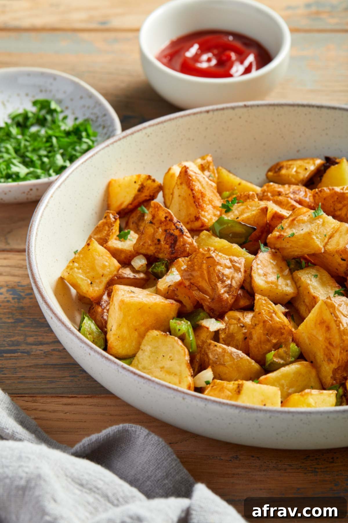Side view of air fryer home fries in a bowl with ketchup and chopped herbs in the background.