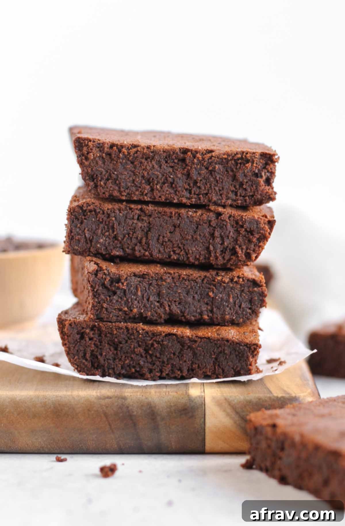 A stack of four fudgy oat flour brownies on a wooden cutting board with a bowl of chocolate chips.