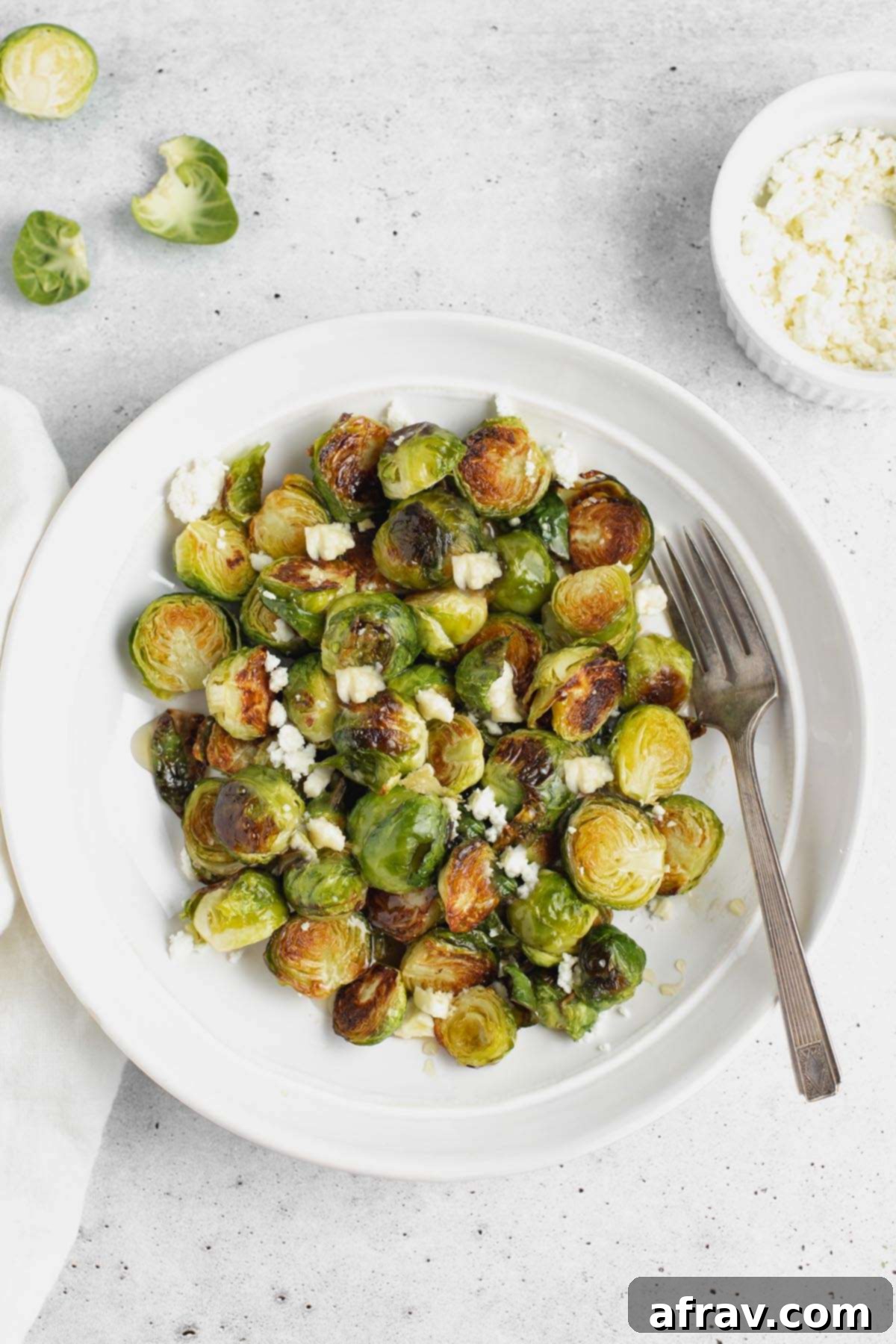 Roasted Brussels sprouts with goat cheese on a round plate with a vintage fork, with raw Brussels in the background on a wooden board.