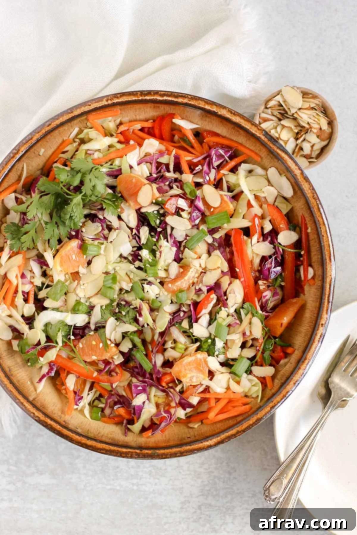 Asian salad in a large ceramic bowl with a small dish of almond slices and stacked plates on the side.