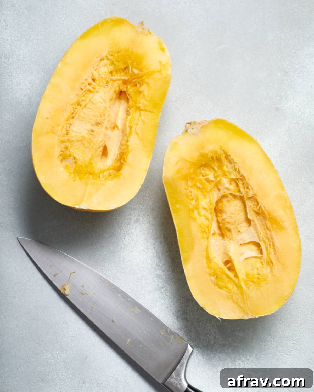 A spaghetti squash carefully cut in half lengthwise, positioned next to a chef's knife on a cutting board, demonstrating the initial preparation step.