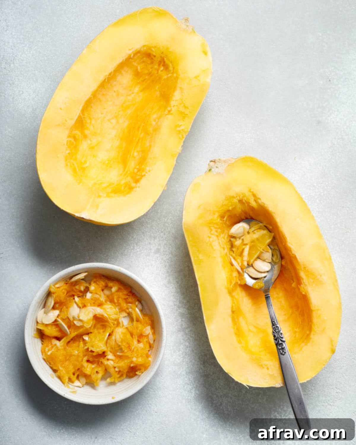 Seeds being meticulously scooped out from one half of a spaghetti squash with a spoon and collected in a small bowl, showcasing the seed removal process.