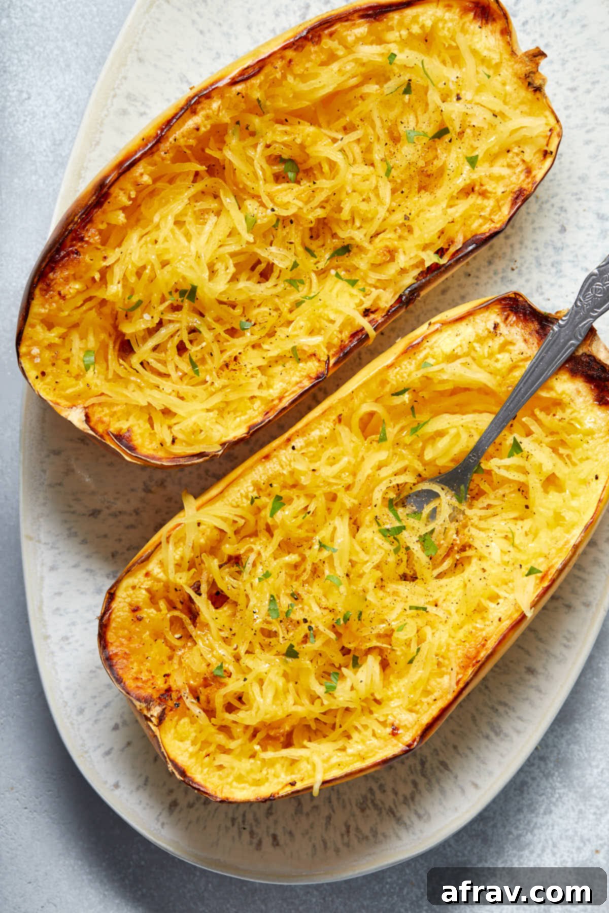 A close-up view of two perfectly roasted spaghetti squash halves on a white platter, with some 'noodles' elegantly twirled around a fork, ready to be eaten.