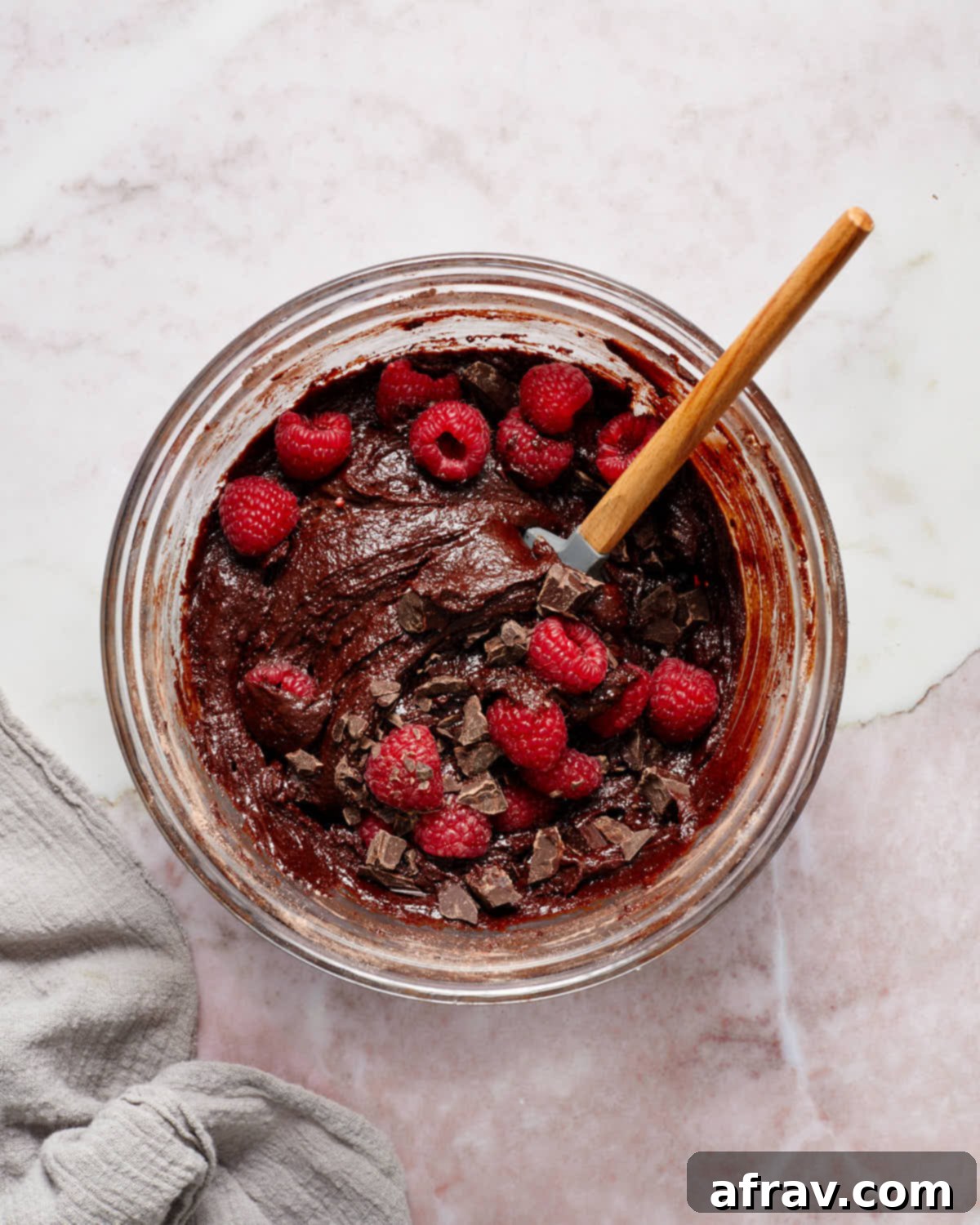 Brownie batter in a glass bowl with raspberries and chopped chocolate stirred in, ready for baking.