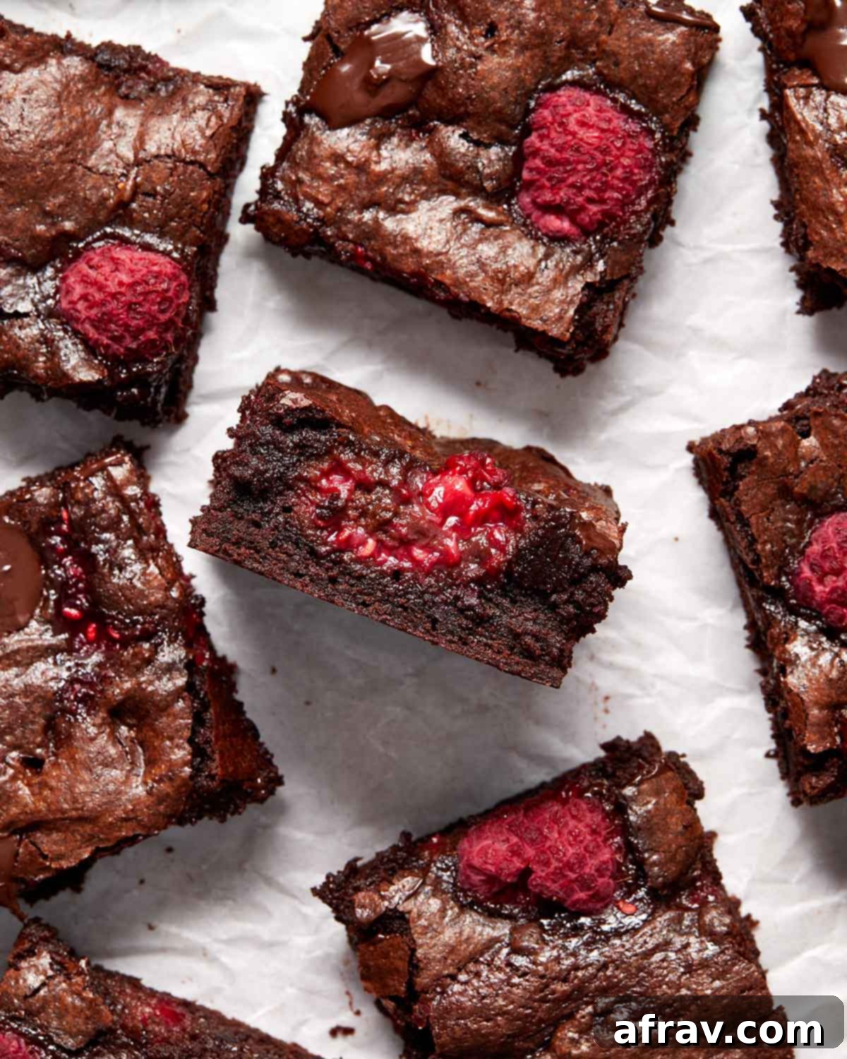 Brownies on white parchment paper with one brownie turned on its side to show the fudgy interior and visible raspberry pieces.