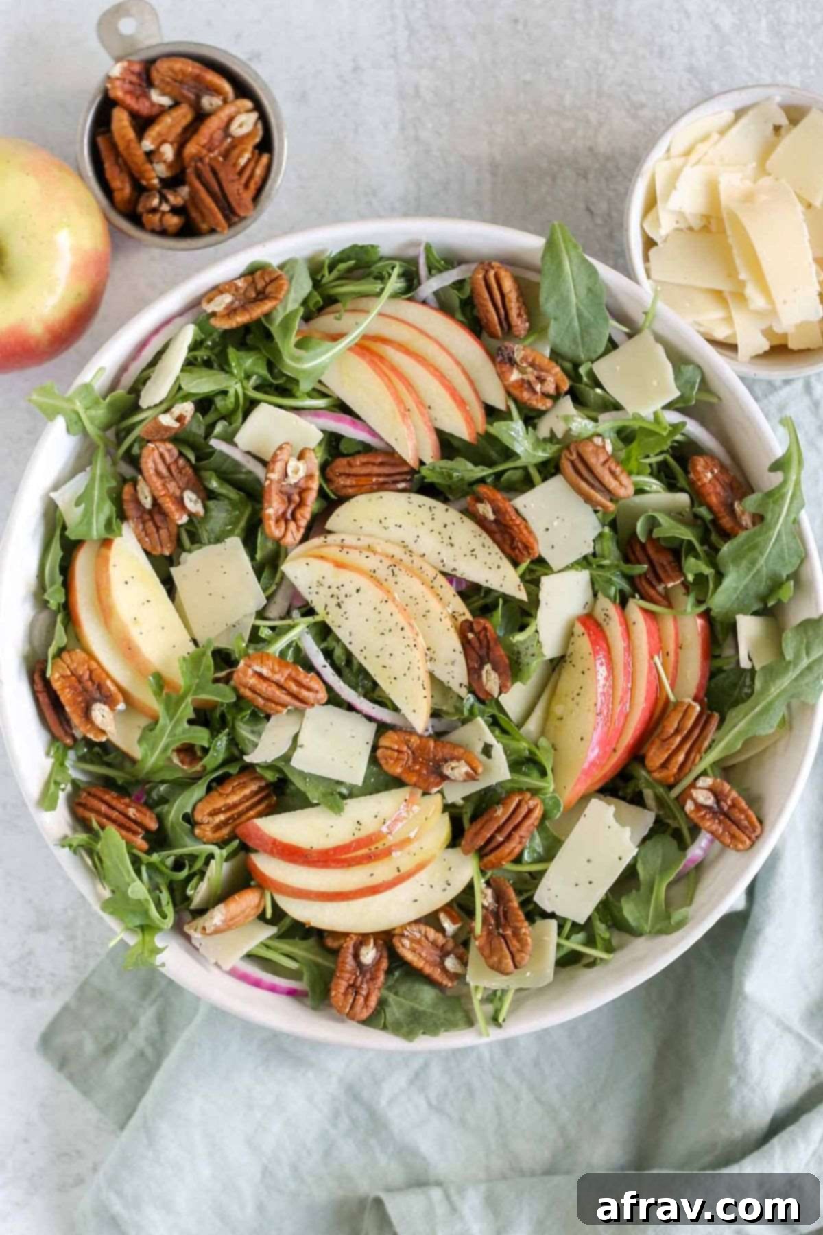 Apple arugula salad in a large serving bowl with sides of parmesan cheese, pecans, and an apple.