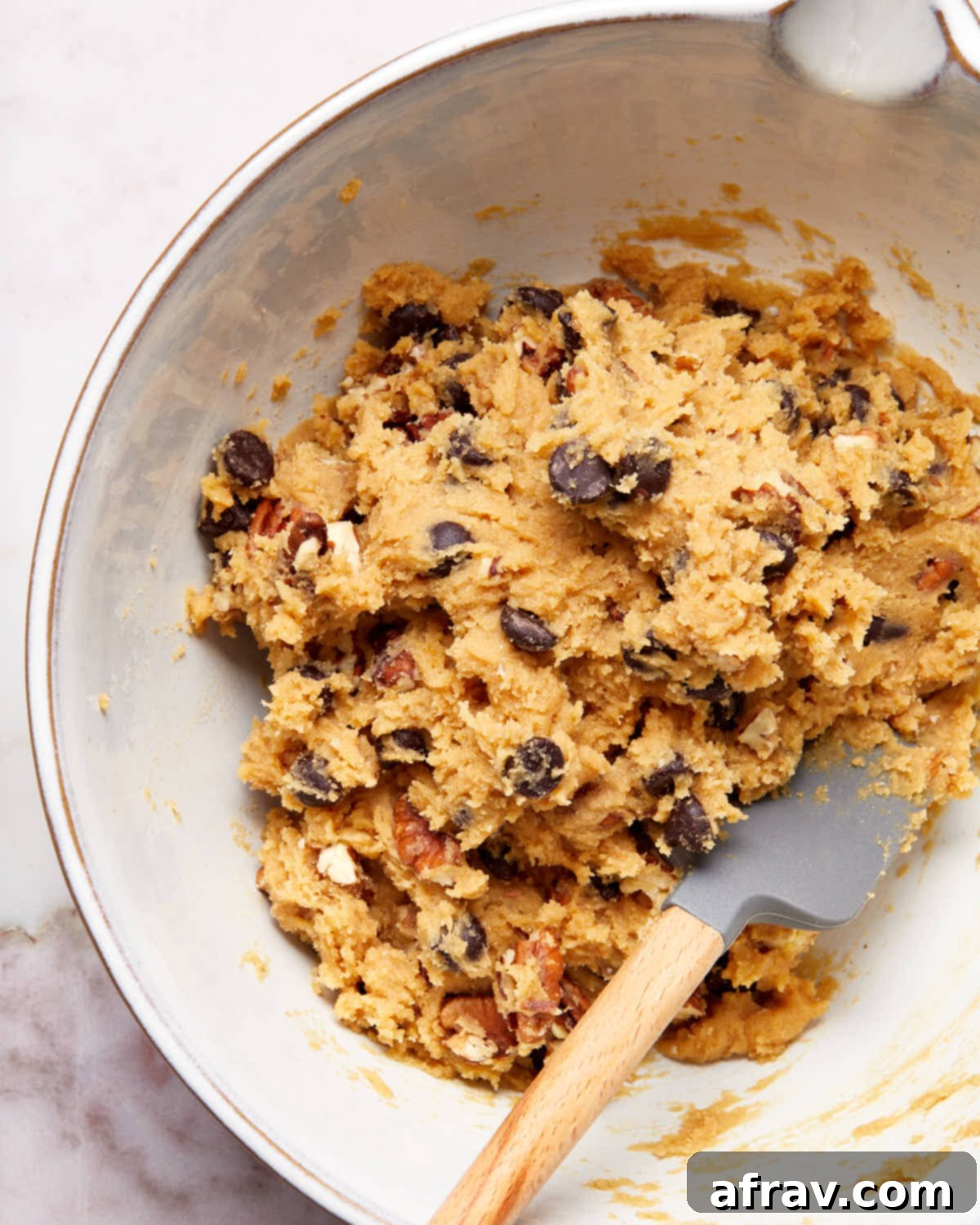 Cookie dough in a white bowl with a spatula inserted into it, showing the mixed chocolate chips and pecans.