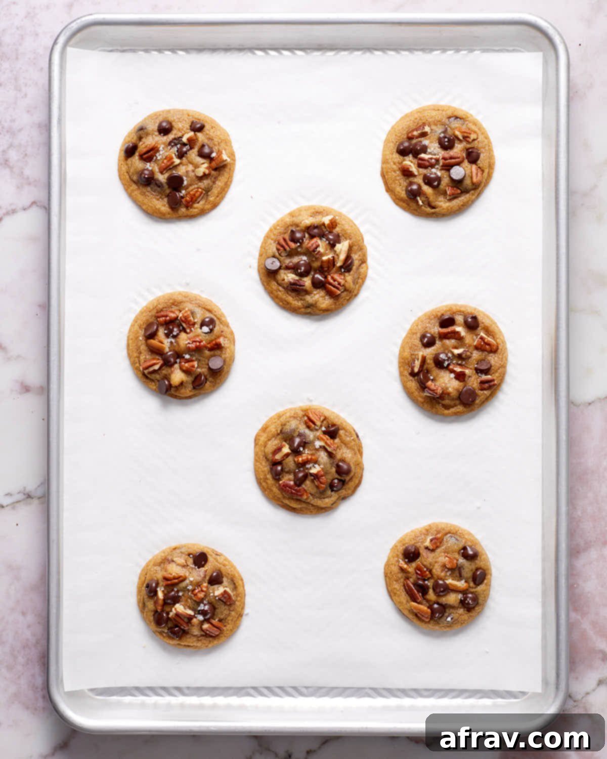 Freshly baked chocolate chip pecan cookies on a baking sheet, glistening with melted chocolate.