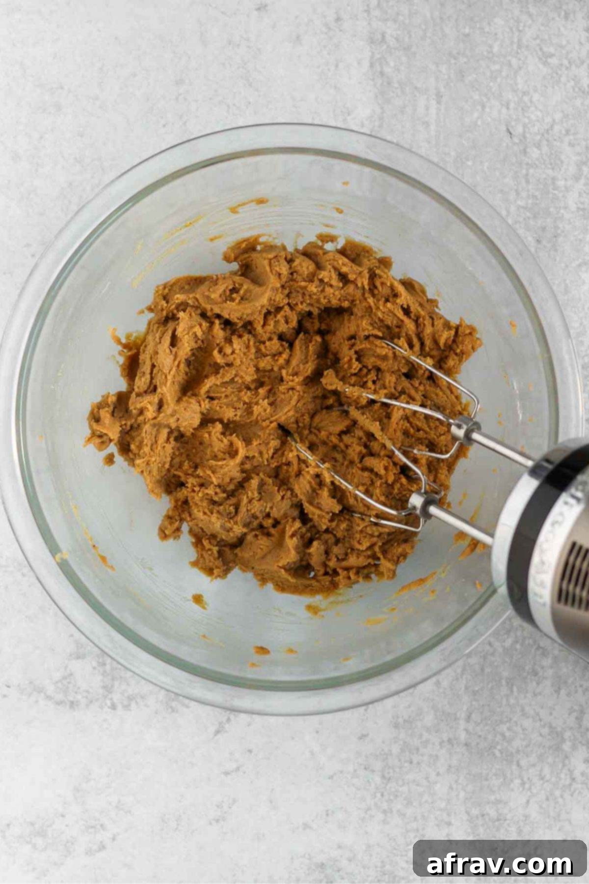 Apple blondie dough being mixed with a hand-mixer in a glass mixing bowl, incorporating dry into wet ingredients.