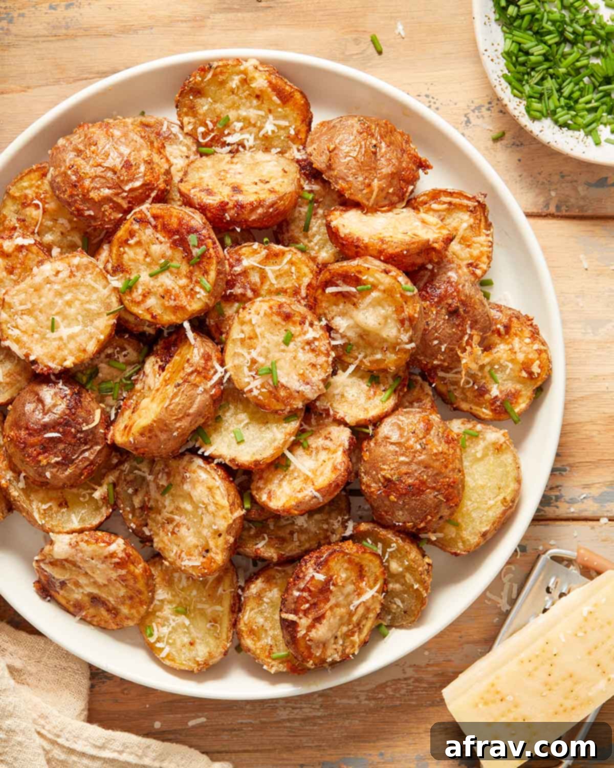 Overhead view of crispy parmesan potatoes on a white plate next to a block of parmesan cheese, chopped chives and a napkin.