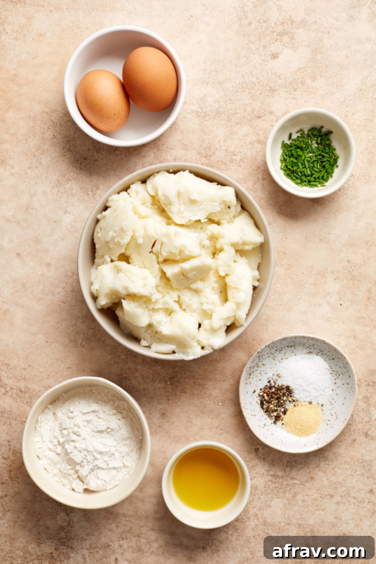 Arrangement of ingredients in individual bowls for making air fryer potato cakes, including mashed potatoes, eggs, flour, seasonings, and chives.